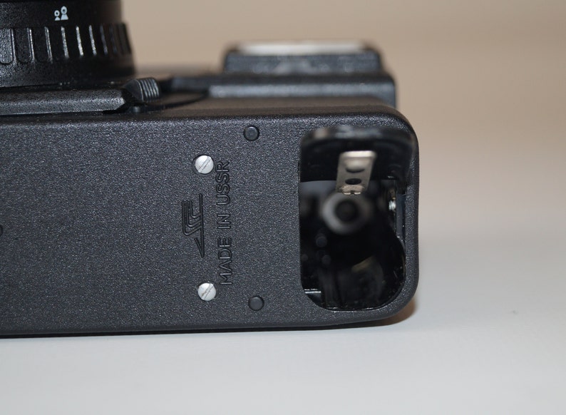 May include: Close-up of a black camera body with a lens and open battery compartment. The camera has a textured surface and the text "MADE IN USSR" is visible. The camera is on a white surface.