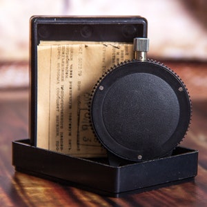 May include: A black, circular timer with a textured dial and a silver winding knob, set in a black rectangular case. The case contains a folded paper with text. The timer is on a wooden surface.