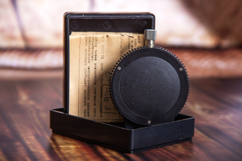 May include: A black, circular timer with a textured dial and a silver winding knob, set in a black rectangular case. The case contains a folded paper with text. The timer is on a wooden surface.