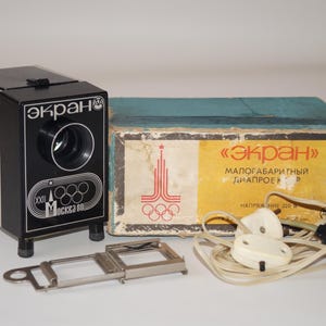 Puede incluir: Proyector de diapositivas vintage en blanco y negro, con los anillos olímpicos y el texto "MOCKBA 80". Incluye un soporte para diapositivas, una caja con texto cirílico y un cable de alimentación. Artículo retro.