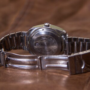 May include: A vintage silver-toned wristwatch with a metal band, resting on a brown leather surface. The watch's back is visible, displaying text and a logo. The band is open, revealing the clasp. The watch appears to be an older model.