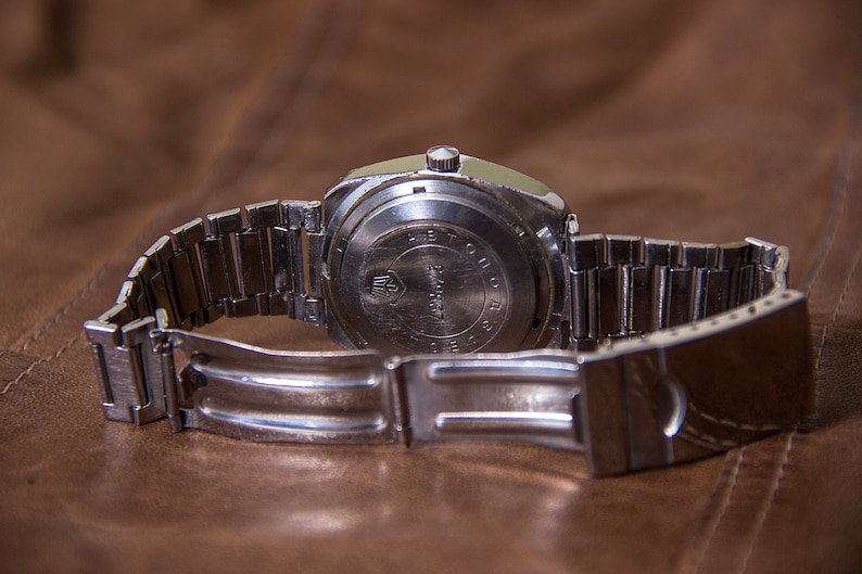 May include: A vintage silver-toned wristwatch with a metal band, resting on a brown leather surface. The watch's back is visible, displaying text and a logo. The band is open, revealing the clasp. The watch appears to be an older model.
