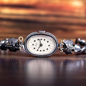 May include: An oval-shaped wristwatch with a textured cream-colored dial, black hands, and black dot hour markers. The watch features a silver-toned metal case and a twisted metal link bracelet with decorative silver and gold-toned accents.