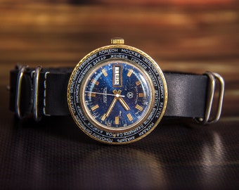 May include: A vintage wristwatch with a gold-tone bezel and a dark blue face. The watch features a black leather band with white stitching and silver-tone hardware. The face displays the time and various city names.