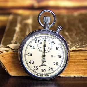 Puede incluir: Un cronómetro plateado con esfera blanca, números negros y un pequeño dial de segundos. Descansa sobre un libro abierto y desgastado con páginas amarillentas. El texto "Агат" es visible en la esfera.