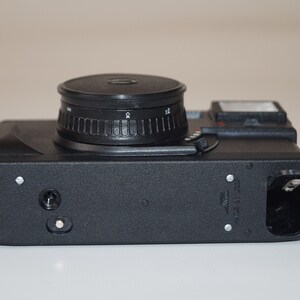 May include: A black film camera with a prominent lens and a flash. The camera body is rectangular with a textured surface. The lens has a ridged adjustment ring. The camera is designed for capturing photographs.