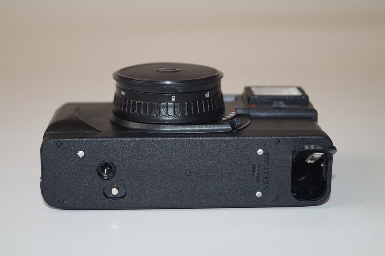 May include: A black film camera with a prominent lens and a flash. The camera body is rectangular with a textured surface. The lens has a ridged adjustment ring. The camera is designed for capturing photographs.