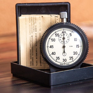May include: A vintage stopwatch with a black casing and a white face, displaying numbers and markings. The stopwatch is in a black box with papers. The text "MADE IN USSR" is visible on the face.