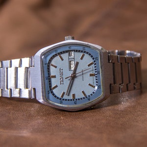 Puede incluir: Un reloj de pulsera plateado vintage con esfera azul claro y correa metálica a juego. La esfera muestra el día y la fecha, junto con la palabra "POLET" en cirílico. El reloj está sobre una superficie de cuero marrón.