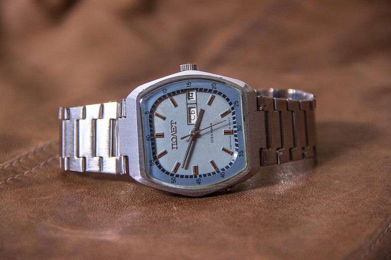 May include: A vintage silver-toned wristwatch with a light blue face and a matching metal band. The watch face displays the day and date, along with the word "POLET" in Cyrillic script. The watch is set against a brown leather surface.