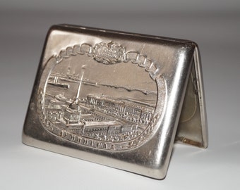 May include: A silver rectangular cigarette case with an embossed cityscape design. The case is open, revealing the interior. The embossed design features buildings, a river, and the word "ЛЕНИНГРАД".