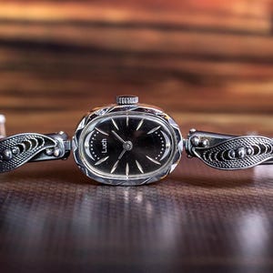 May include: A silver-tone Luch wristwatch featuring an oval-shaped case with a black dial and silver hands. The watch band is composed of intricate filigree metal links on both sides, showcasing a detailed, openwork leaf-like pattern.