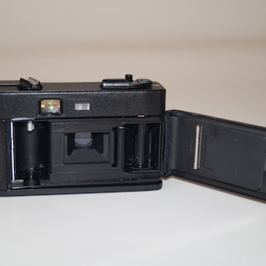 May include: Black vintage film camera with the back open, revealing the interior components. The camera has a flash and a top-mounted shutter button. The open back shows the film compartment and the camera's internal mechanisms.