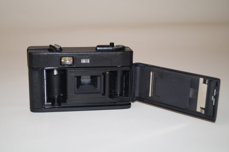 May include: Black vintage film camera with the back open, revealing the interior components. The camera has a flash and a top-mounted shutter button. The open back shows the film compartment and the camera's internal mechanisms.