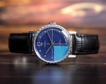 Puede incluir: Un reloj de pulsera vintage con una esfera azul con un patrón texturizado y una sección de cuadrados azul claro. El reloj tiene una caja plateada, correas de cuero negro y el número 12 es visible. La palabra "ПОБЕДА" también es visible.