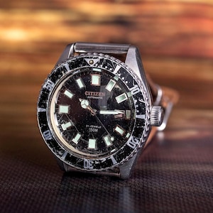 Puede incluir: Un reloj autom&aacute;tico Citizen vintage con esfera negra y marcadores luminosos. El reloj tiene una caja de acero inoxidable y un bisel giratorio con marcas blancas. El reloj tiene una correa de cuero marr&oacute;n. La esfera del reloj dice "Citizen Automatic" y "21 Jewels 150M".