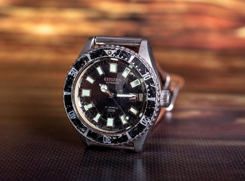Puede incluir: Un reloj autom&aacute;tico Citizen vintage con esfera negra y marcadores luminosos. El reloj tiene una caja de acero inoxidable y un bisel giratorio con marcas blancas. El reloj tiene una correa de cuero marr&oacute;n. La esfera del reloj dice "Citizen Automatic" y "21 Jewels 150M".