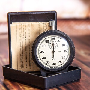 May include: A vintage black stopwatch with a white face, featuring black numerals and markings. The stopwatch is in a black case with a paper insert. The watch measures time in seconds and minutes. The text on the watch face is clear and readable.