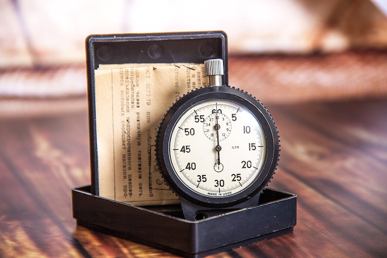 May include: A vintage black stopwatch with a white face, featuring black numerals and markings. The stopwatch is in a black case with a paper insert. The watch measures time in seconds and minutes. The text on the watch face is clear and readable.