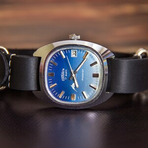 May include: A vintage Cornavin watch with a blue gradient face and a black leather band. The watch has a silver-tone case and a date display at the 3 o'clock position. The band is approximately 20 mm wide.