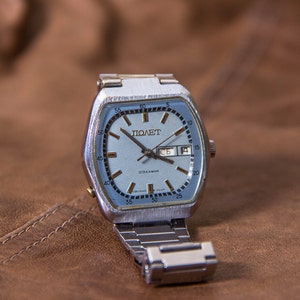 May include: A vintage silver-toned wristwatch with a light blue face and a date display. The watch face features the word "ПОЛЕТ" and other Cyrillic text. The watch has a silver metal band and is set against a brown leather background.