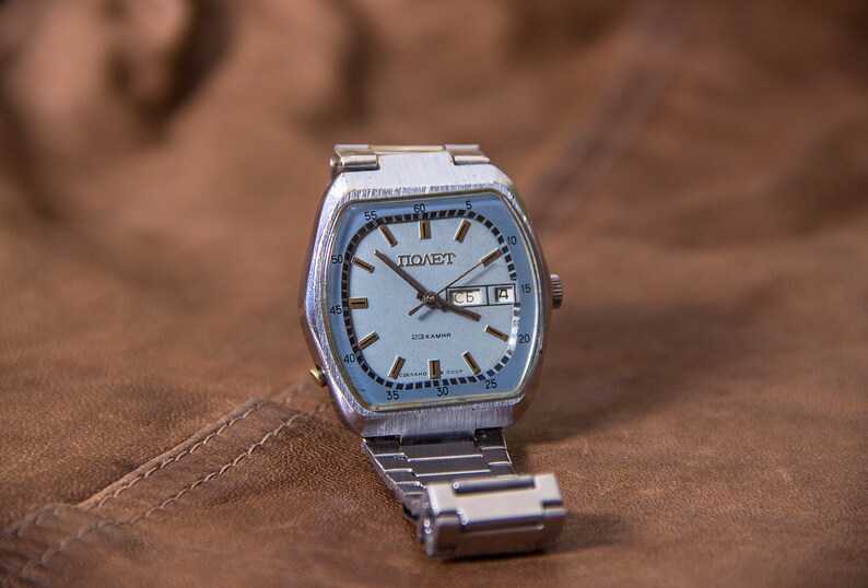May include: A vintage silver-toned wristwatch with a light blue face and a date display. The watch face features the word "ПОЛЕТ" and other Cyrillic text. The watch has a silver metal band and is set against a brown leather background.