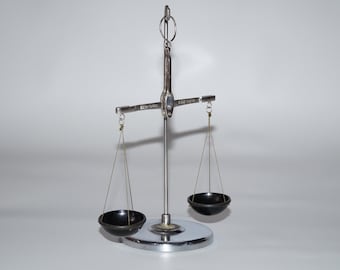 Puede incluir: Una balanza de metal plateado con un poste vertical central, una barra horizontal y dos cuencos negros pequeños suspendidos por hilos finos. La balanza descansa sobre una base circular plateada e incluye un anillo en la parte superior.