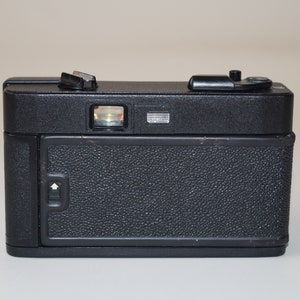 May include: A black vintage camera with a textured body. The camera has a viewfinder, flash mount, and various dials and buttons on top. The back of the camera features a textured door with a latch. The camera is a classic piece of photography equipment.
