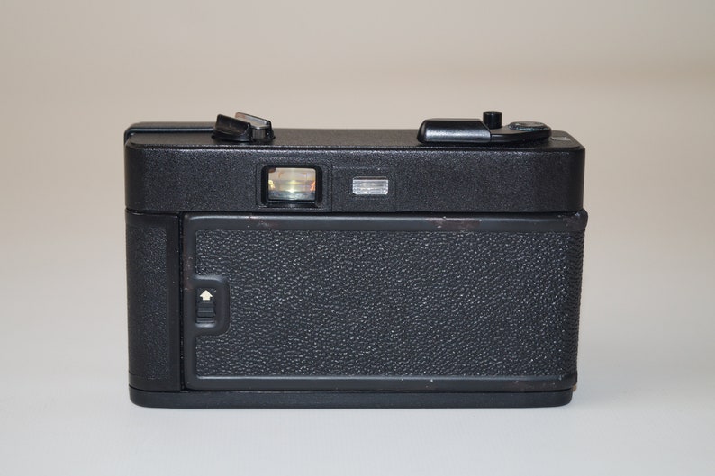 May include: A black vintage camera with a textured body. The camera has a viewfinder, flash mount, and various dials and buttons on top. The back of the camera features a textured door with a latch. The camera is a classic piece of photography equipment.