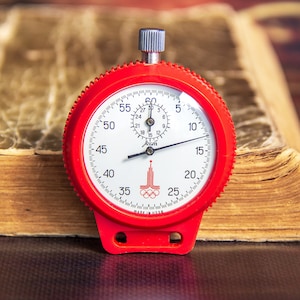 May include: A red stopwatch with a white face and black numerals sits on an open, aged book. The stopwatch has a silver winding knob and a logo with Olympic rings. The book's pages are yellowed, suggesting age.