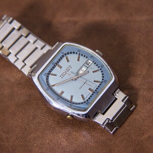 May include: A vintage silver-toned wristwatch with a light blue face and a date display. The watch has a square face with rounded corners and a silver metal band. The watch face has the word "ПОЛЕТ" printed on it.