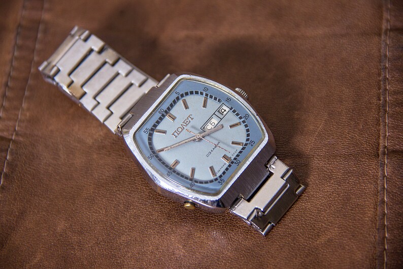 May include: A vintage silver-toned wristwatch with a light blue face and a date display. The watch has a square face with rounded corners and a silver metal band. The watch face has the word "ПОЛЕТ" printed on it.