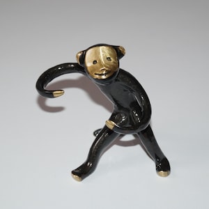 Puede incluir: Pequeña figura de mono negro con cara, orejas y detalles dorados. El mono está posando de forma juguetona, con un brazo extendido y el otro doblado. La figura está sobre un fondo blanco liso.