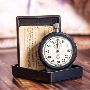 May include: A vintage stopwatch with a black casing and a white face, displaying numbers and markings. The stopwatch is in a black box with a paper document. The background is a wooden surface.