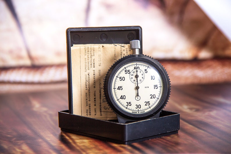 May include: A vintage stopwatch with a black casing and a white face, displaying numbers and markings. The stopwatch is in a black box with a paper document. The background is a wooden surface.