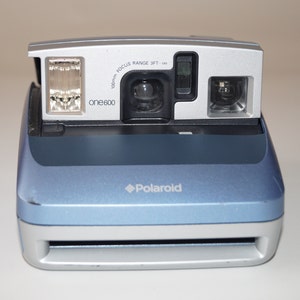 Peut inclure: Un appareil photo instantané Polaroid One600 bleu et argenté. L'appareil photo est équipé d'un flash intégré, d'un objectif et d'un viseur. Le logo Polaroid est visible à l'avant. La plage de mise au point est de 0,91 mètre à l'infini.