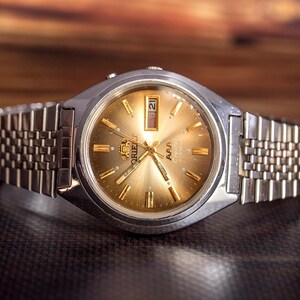 Può includere: Un orologio da polso color argento con quadrante dorato a raggiera, dotato di indici e lancette color oro. L'orologio include una finestrella giorno-data a ore tre, il logo Orient e un cinturino in metallo color argento a maglie multiple.
