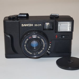 May include: Black vintage camera with the text "Зенит 35CM" and "ИНДУСТАР-95 2,8/38" on the lens. The camera has a flash and a lens cap. The camera is a classic film camera.