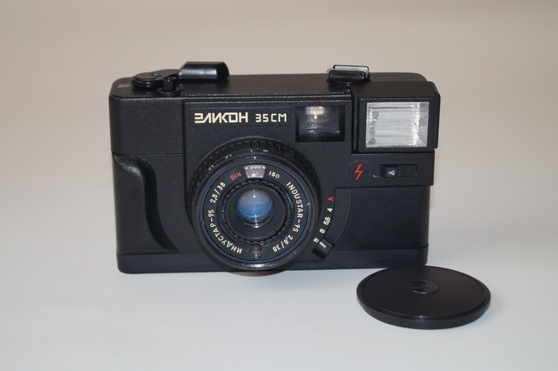 May include: Black vintage camera with the text "Зенит 35CM" and "ИНДУСТАР-95 2,8/38" on the lens. The camera has a flash and a lens cap. The camera is a classic film camera.