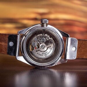 Puede incluir: Primer plano de un reloj Citizen vintage, que muestra los intrincados mecanismos internos a trav&eacute;s de una parte trasera transparente. El reloj tiene una caja plateada y una correa de cuero marr&oacute;n. La esfera dice "Citizen Japan 21 Jewels".