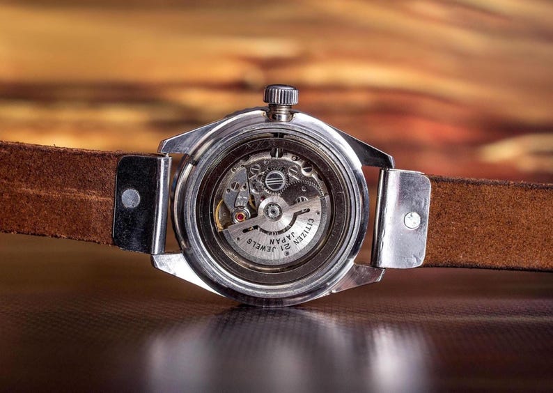 Puede incluir: Primer plano de un reloj Citizen vintage, que muestra los intrincados mecanismos internos a trav&eacute;s de una parte trasera transparente. El reloj tiene una caja plateada y una correa de cuero marr&oacute;n. La esfera dice "Citizen Japan 21 Jewels".