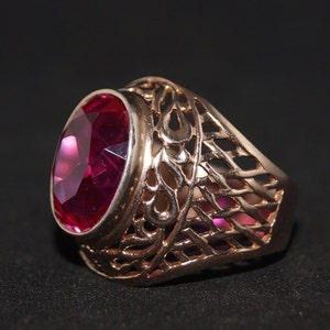 May include: A rose gold ring with an oval, faceted, deep red gemstone. The ring features an open filigree design on the band, with a dark background.