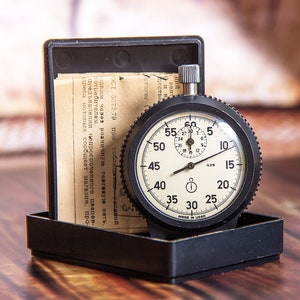May include: A vintage stopwatch with a black casing and a cream-colored face, displaying numbers and markings. The stopwatch is in a black box with paperwork. The watch reads "MADE IN USSR".