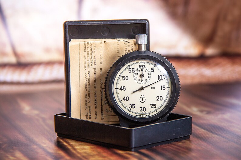 May include: A vintage stopwatch with a black casing and a cream-colored face, displaying numbers and markings. The stopwatch is in a black box with paperwork. The watch reads "MADE IN USSR".