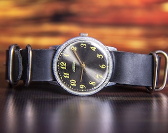 May include: A vintage wristwatch with a black face and gold hands and numerals. The watch has a silver-colored bezel and is attached to a black leather strap. The watch face has the word "Tocka" printed on it.