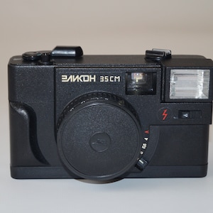 May include: A black "Elikon 35CM" camera with a lens, flash, and various dials. The camera has a rectangular body with rounded edges. The text "Elikon 35CM" is printed in white on the front of the camera.