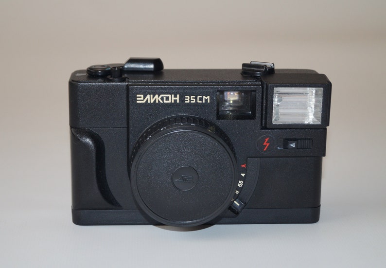 May include: A black "Elikon 35CM" camera with a lens, flash, and various dials. The camera has a rectangular body with rounded edges. The text "Elikon 35CM" is printed in white on the front of the camera.