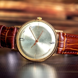 May include: A gold-toned wristwatch with a silver face and brown leather band. The watch face features black numerals at 12 and 6 o'clock, with the brand name "Luch" in script. The band is textured with a crocodile pattern.