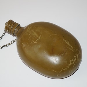 May include: Olive green canteen with a screw-on cap and a metal chain. The canteen has an oval shape and a textured surface. The cap is attached to the canteen with a small metal chain.
