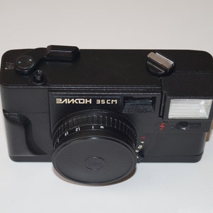 May include: Black Elikon 35CM camera with a lens cap. The camera has a flash and a silver-colored button on top. The text "ЭЛИКОН 35 CM" is printed on the front of the camera. The camera is on a white surface.